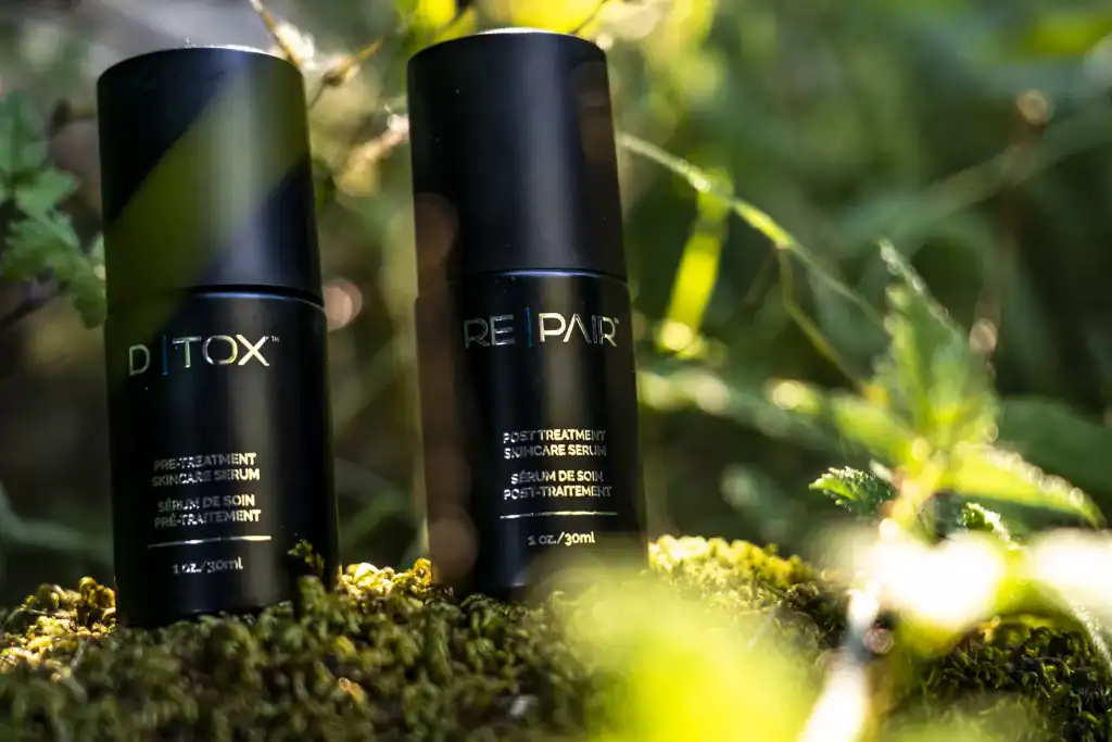 Two black skincare serum bottles on moss with greenery background.
